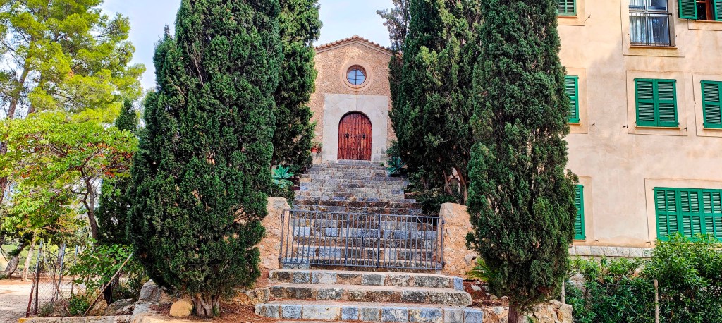 Escalera de piedra con barandilla que lleva a la entrada de la Ermita de Santa Lucía, rodeado de cipreses y vegetación, en un entorno natural.