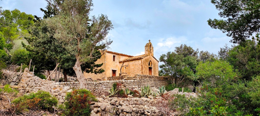 Vista del Oratorio de Son Mascord, una pequeña construcción de piedra rodeada de vegetación y árboles, en un paisaje natural de Mallorca.