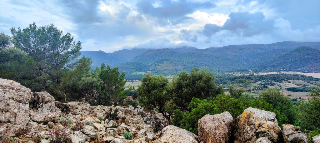 Vista panorámica de un paisaje montañoso y verde, con árboles y rocas, bajo un cielo nublado  que se observa desde el Camí de s’Ullastrar