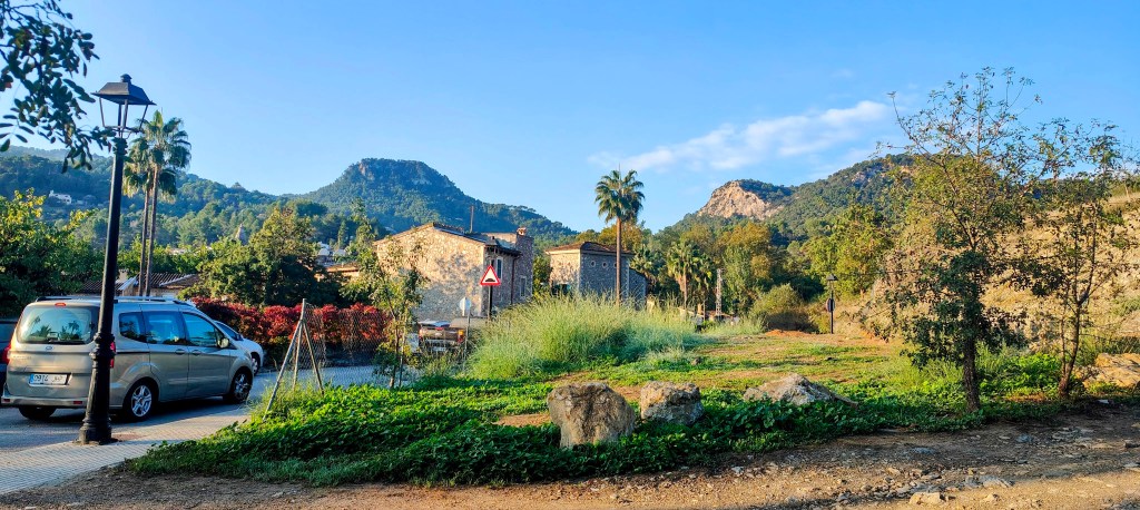 Vista del pueblo de Puigpunyent con montañas de fondo, un coche aparcado y un entorno natural que incluye árboles y senderos.