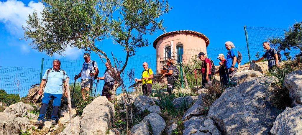 Grupo de senderistas descendiendo por el Camí de sa Cova des Bous por un terreno rocoso en la ruta 'Penyal de Sa Mola', con un edificio en el fondo y un cielo despejado.