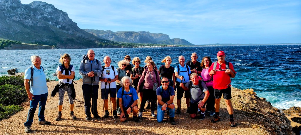 Grupo de caminantes posando en la Punta des Caló con montañas de fondo, en un día soleado.