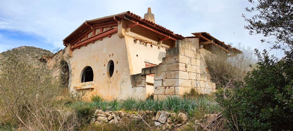 Edificio en ruinas con paredes de color claro y detalles de ladrillo. Rodeado de vegetación y montañas.