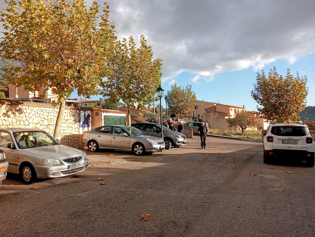 Vista del aparcamiento en Mancor de la Vall, con varios coches estacionados y árboles en el ambiente, junto a edificios locales.