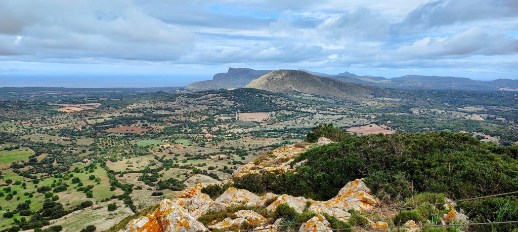Vista panorámica desde la cima del Puig d’en Sard, mostrando un paisaje de colinas y campos en la Serra de Calicant, con nubes en el cielo.