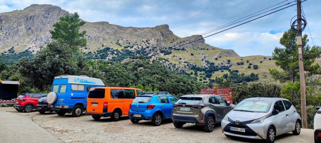 Vista de un aparcamiento con varios coches, rodeado de montañas y vegetación, que sirve como punto de partida para la ruta 'Els Arcs-Lluc-Els Arcs'.