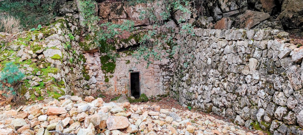 Entrada de la Font d’Escorça una construcción de piedra rodeada de rocas y vegetación, en un área montañosa.