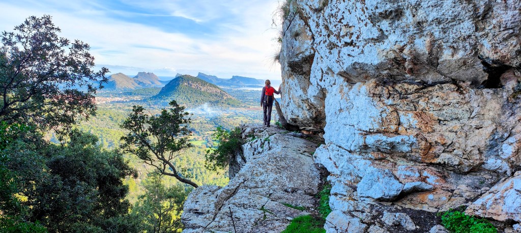 Montañés caminando por un sendero estrecho junto a un acantilado, con vistas panorámicas de un valle y montañas al fondo.