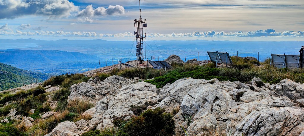 Vista desde la cima de la Tossa del Llamp, mostrando una instalación de telecomunicaciones rodeada de rocas y vegetación, con un paisaje montañoso y nubes en el cielo.