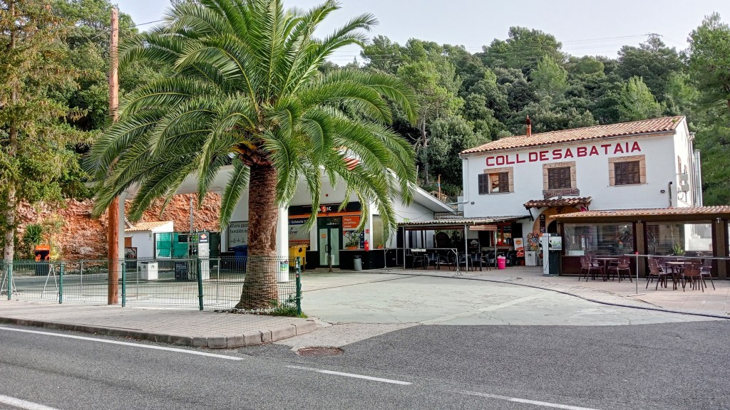 Vista del Coll de Sa Batalla, con una palmera en primer plano, una gasolinera al fondo y un restaurante rústico con el nombre visible en su fachada.