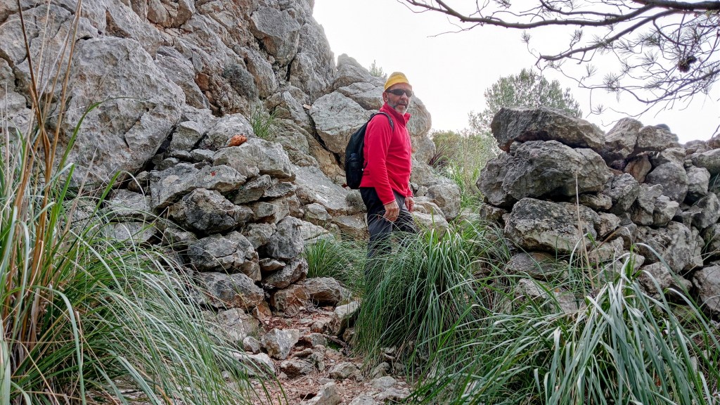 Persona en el Pas d’en Bartomeu rodeado de rocas y hierba, vistiendo una camiseta roja y un gorro amarillo.