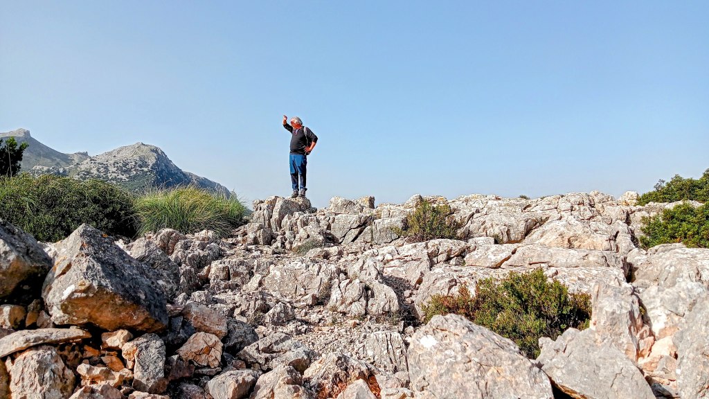 Una persona de pie sobre rocas en la cima del Puig de Ses Covasses, señalando hacia el horizonte con una mano. Al fondo, se pueden ver montañas en un día soleado.