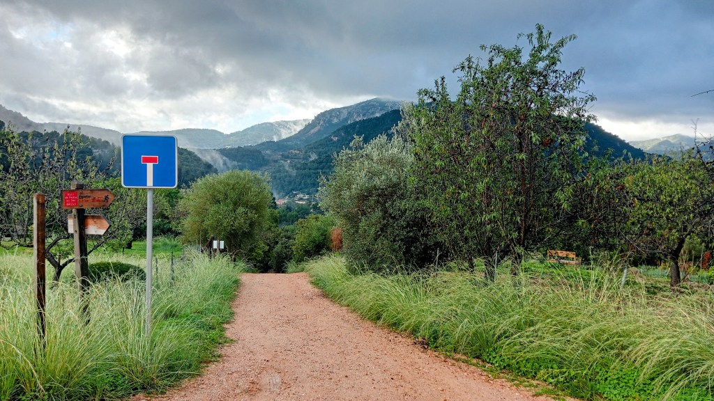 Camino de tierra con señal de tráfico que indica el final de la vía para vehículos, rodeado de vegetación y montañas al fondo.