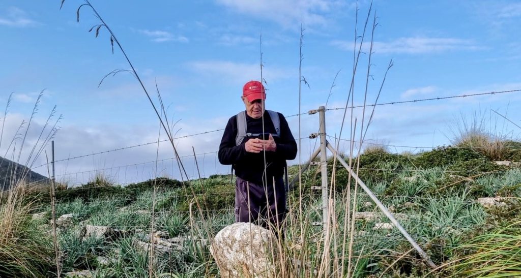 Una persona con una gorra roja revisa su teléfono móvil, rodeado de vegetación y una cerca, bajo un cielo azul con nubes en el Puig d’en Pelat.
