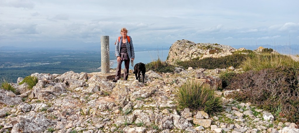 Persona de pie junto a un vértice geodésico en la cima de Es Ferruxet, con un paisaje montañoso y una vista panorámica del valle y el mar al fondo.