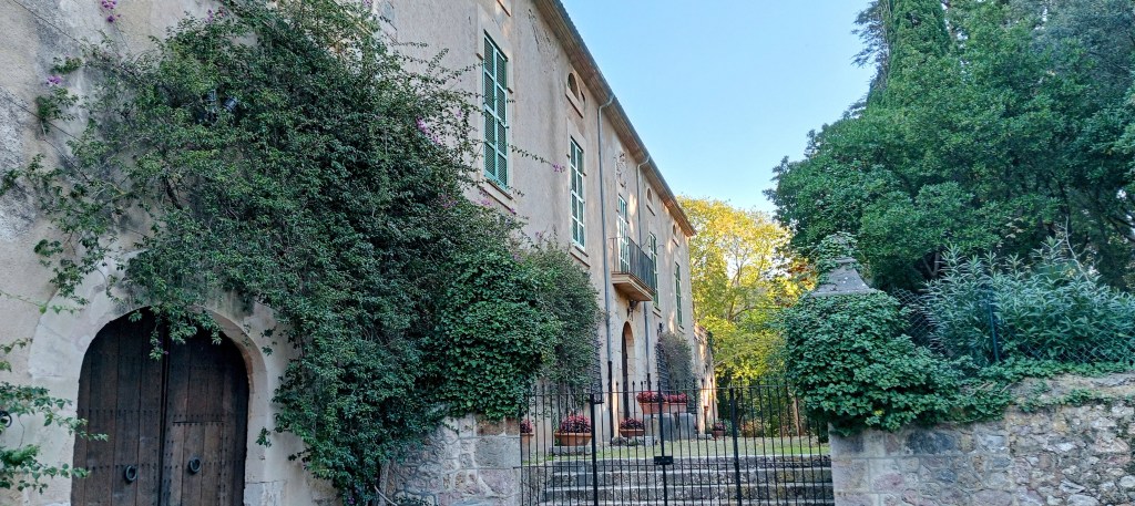 Vista de la fachada de las casas de Son Grúa con puertas de madera, rodeada de vegetación exuberante y un jardín al fondo.