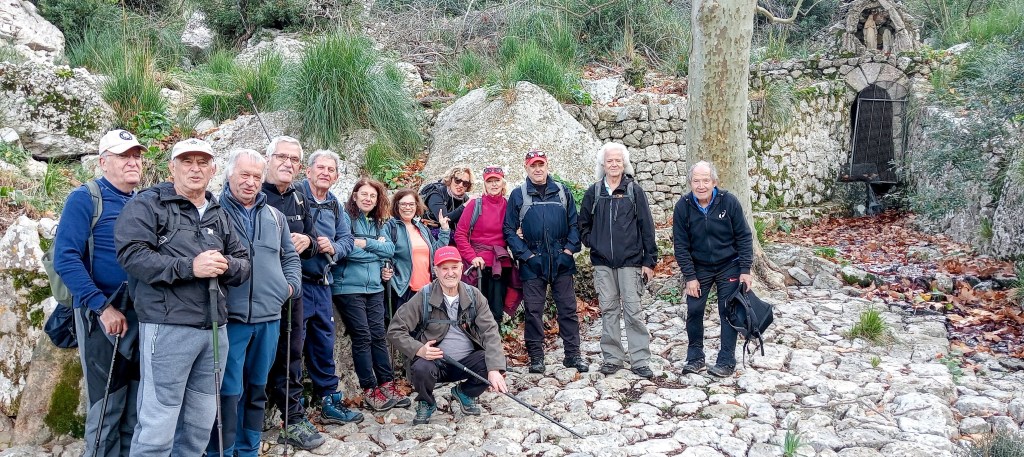 Grupo de excursionistas posando en un sendero rocoso cerca de la Font de la Mare de Déu en un entorno natural, con árboles y vegetación alrededor.