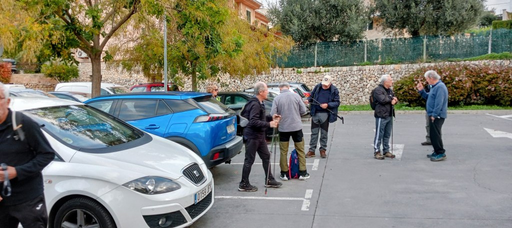 Grupo de senderistas preparándose para iniciar la ruta en el aparcamiento del pueblo de Es Capdellà, rodeado de vehículos y vegetación.