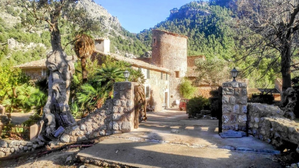 Entrada a una casa de piedra rodeada de montañas y vegetación, con un camino claro y árboles a los lados.