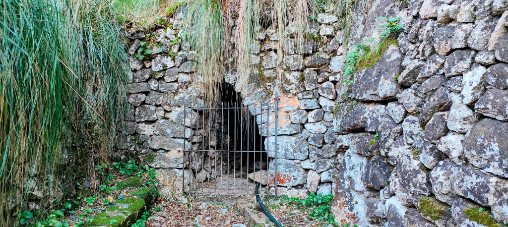 Entrada de la Font de Bàlitx d’Enmig con una reja de metal, rodeada de paredes de piedra y vegetación cerca.
