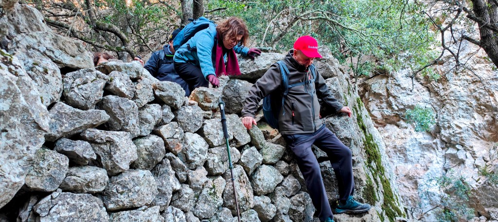 Dos personas escalando un muro de piedras en el Pas Nou , con vegetación detrás.