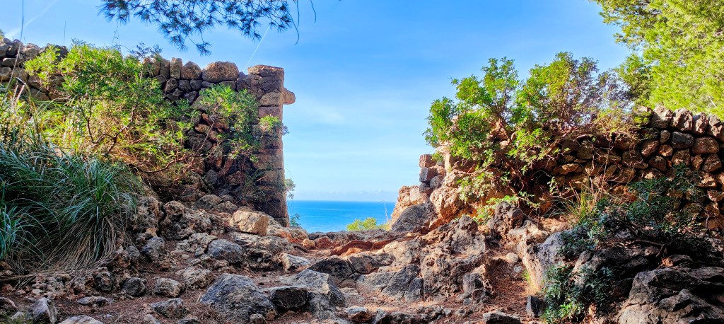 Vista de un camino rocosa rodeada de vegetación y muros de piedra, con el mar al fondo y un cielo azul despejado.