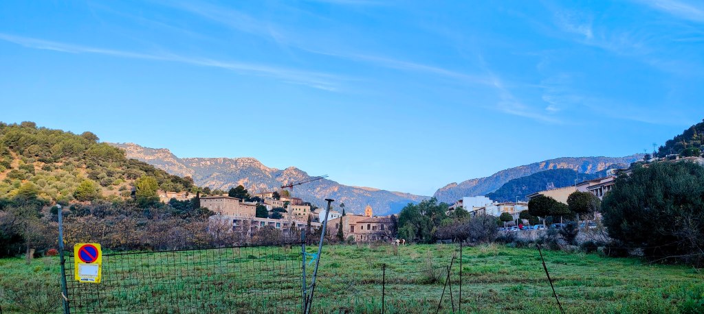 Paisaje rural con montañas al fondo, casas antiguas y un campo verde en primer plano, bajo un cielo azul despejado.