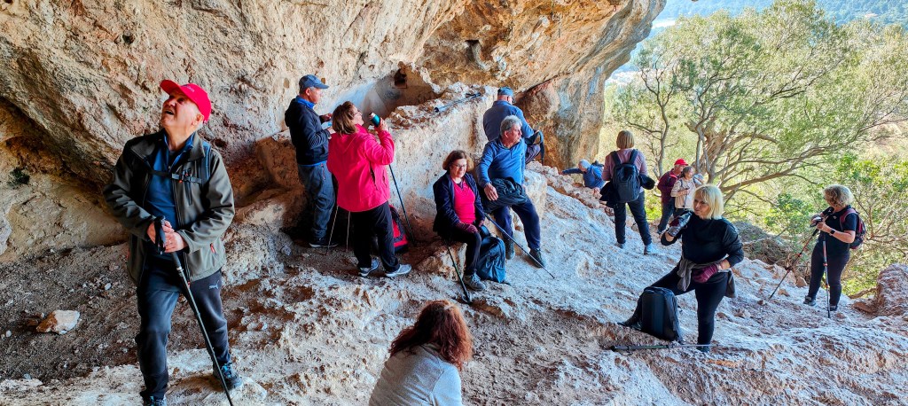 Grupo de personas en un refugio rocoso, algunos de pie y otros sentados, disfrutando de una actividad al aire libre.