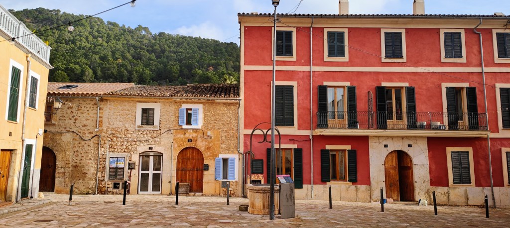 Plaza con arquitectura tradicional, edificios de piedra y colorido, rodeada de vegetación montañosa.