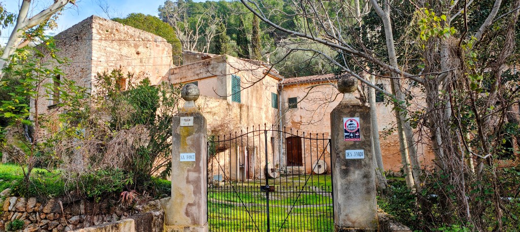 Vista de las casas de sa Font des Jardí rodeada de vegetación, con una entrada enrejada y columnas decorativas a los lados.