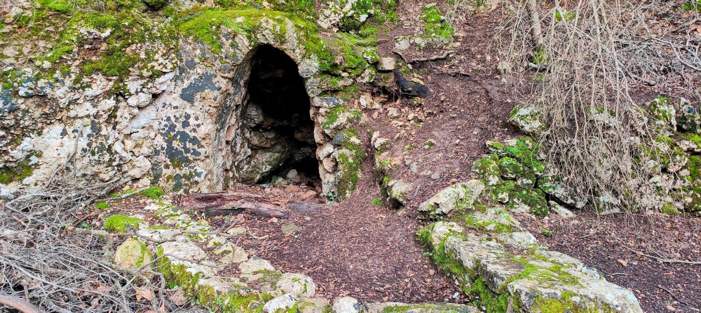 Entrada a la Font des Freu entre rocas cubiertas de musgo y tierra en un entorno natural.