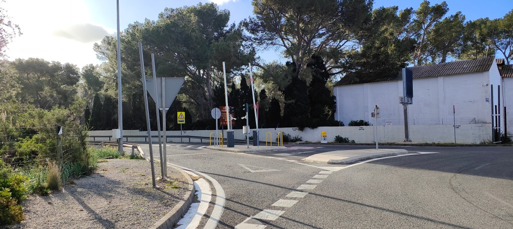 carretera de Formentor rodeada de árboles con señales de tráfico y un edificio blanco al fondo.