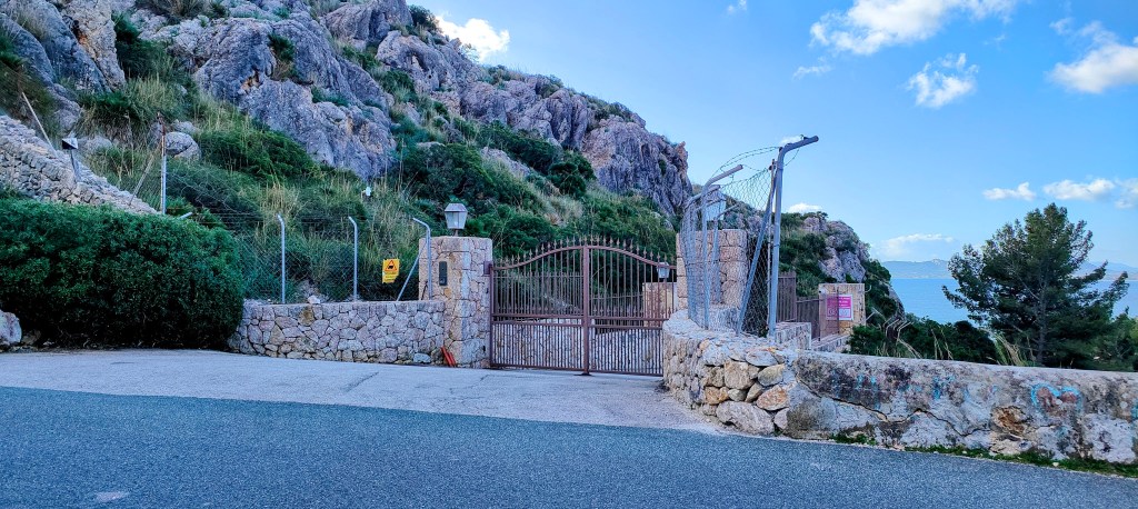 Puerta de hierro rodeada de piedras y vegetación, con vistas al mar y montañas al fondo.