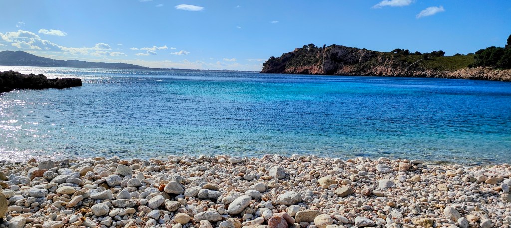 Vista de la Cala "Es Caló" de piedras con agua azul clara y un paisaje costero al fondo bajo un cielo parcialmente nublado.