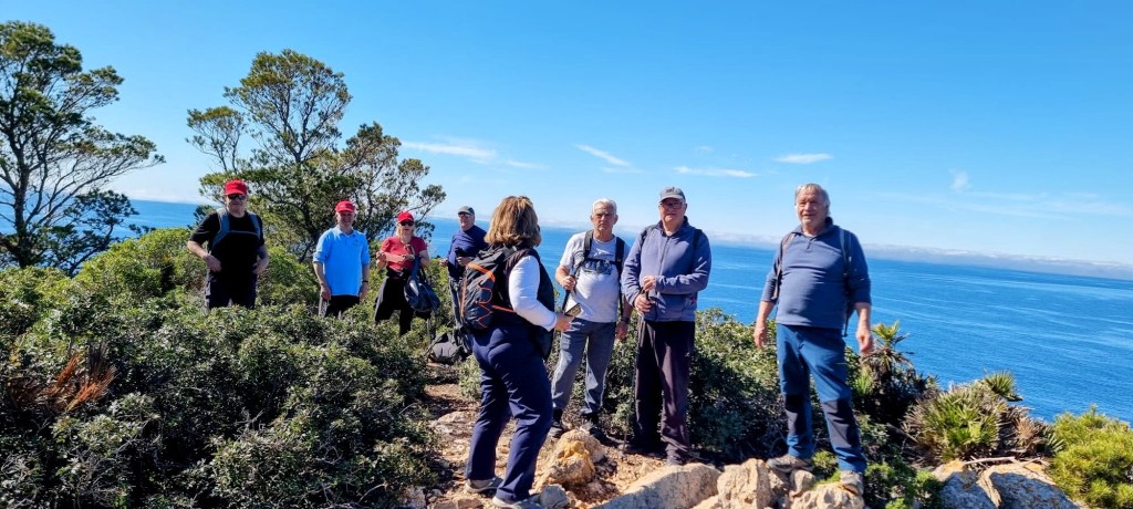 Un grupo de ocho personas en una cima montañosa, disfrutando de una vista al mar con un cielo despejado.