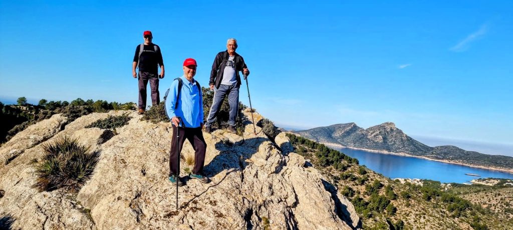 Tres excursionistas en la cima rocosa de Es Tres Picons con vistas a un paisaje montañoso y un lago en el fondo, bajo un cielo despejado.