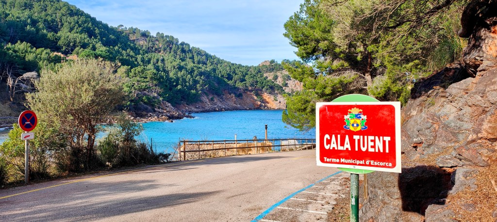 Vista de la Cala Tuent, con un cartel que indica la ubicación en el municipio de Escorca, rodeada de montañas y vegetación.