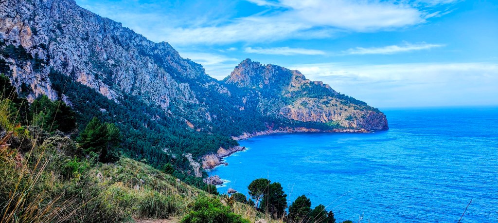 Vista panorámica de un acantilado junto al mar, con montañas y vegetación en primer plano bajo un cielo azul.