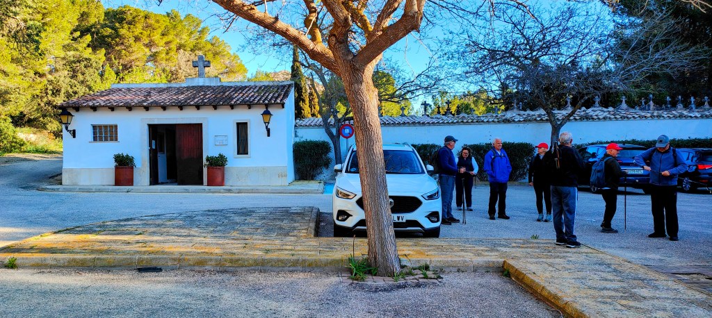 Un grupo de personas rodea un coche blanco frente a una pequeña capilla con una cruz en el techo, en un entorno natural con árboles.