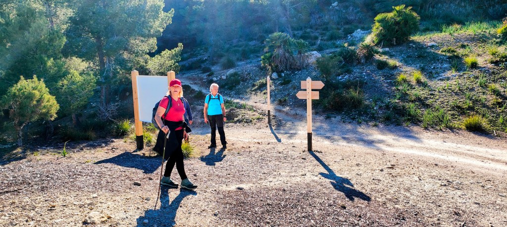 Dos senderistas de pie en un camino rural con señalización, rodeados de naturaleza y árboles.