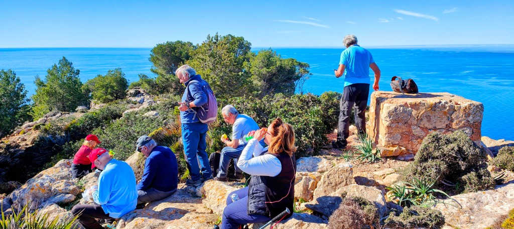 Grupo de personas disfrutando de un descanso en la cima del Puig d’en Tió con vista al mar, rodeados de vegetación.