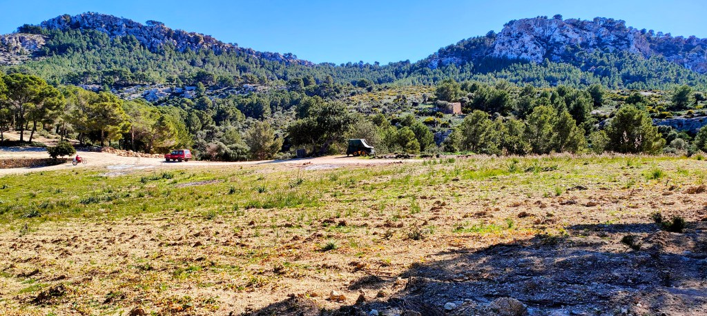 Vista de un paisaje montañoso con vegetación y un terreno despejado. Se observa un vehículo rojo y una pequeña construcción al fondo.