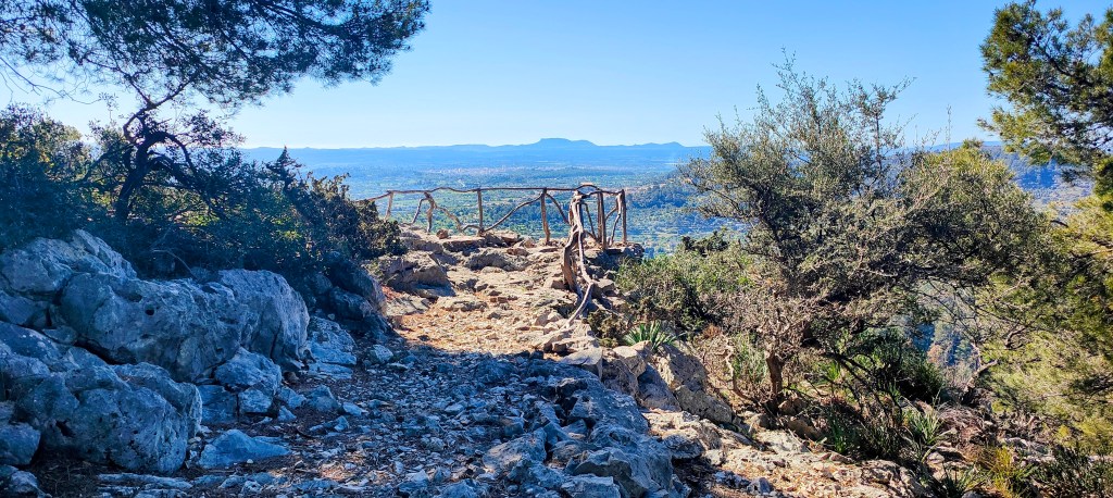 Sendero rocoso que conduce a un mirador con vista panorámica al valle y montañas, rodeado de vegetación.