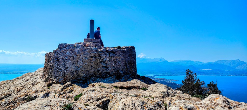 Un hombre sentado en la cima de una roca, mirando el paisaje montañoso y el mar al fondo, con un cielo claro y soleado.