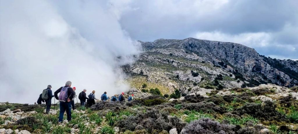 Grupo de personas caminando por un sendero montañoso, rodeados de nubes y vegetación, con formaciones rocosas al fondo.