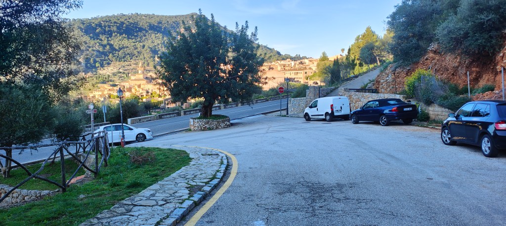 Vista de una carretera en una zona rural, con árboles y casas en la colina. Se observan vehículos estacionados en la calzada.