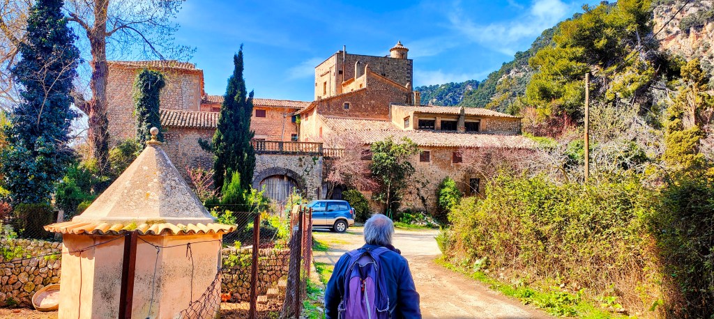 Una persona caminando hacia un antiguo edificio de piedra en un entorno rural, rodeado de árboles y vegetación. Se puede ver un coche aparcado y un pequeño torreón al fondo.