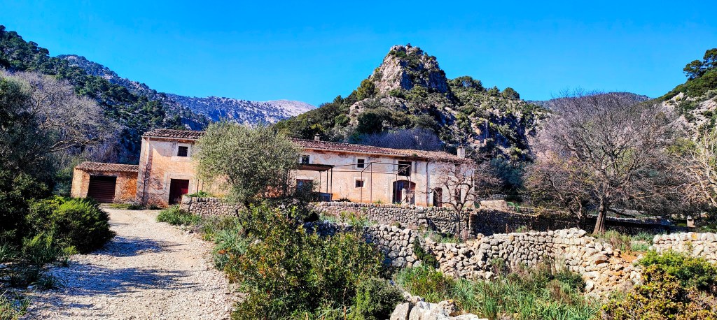 Vista de una casa rural rodeada de naturaleza, con montañas al fondo y un camino de gravilla que lleva hasta la entrada.