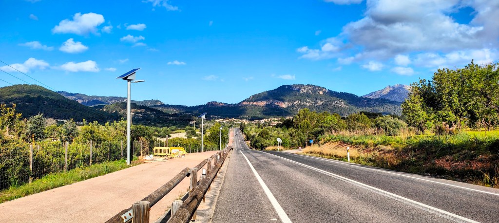 Carretera rural rodeada de montañas y vegetación bajo un cielo azul con algunas nubes.