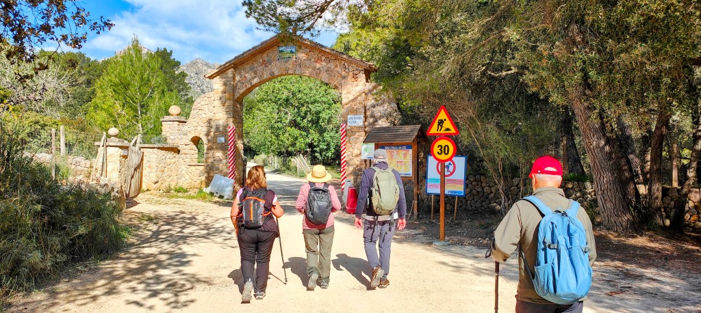 Cuatro personas caminando por un sendero hacia un arco de piedra, rodeado de árboles y señales de tráfico.